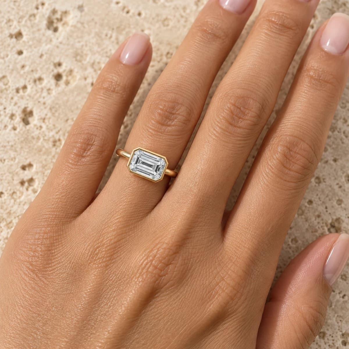 Emerald-cut diamond set east-west, lying across the finger in a yellow gold bezel on a plain polished solitaire band, worn on a woman's left ring finger against warm travertine