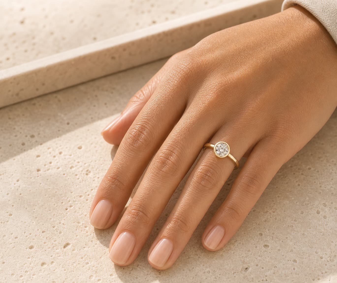 Round bezel engagement ring on a woman's left hand ring finger against warm travertine stone